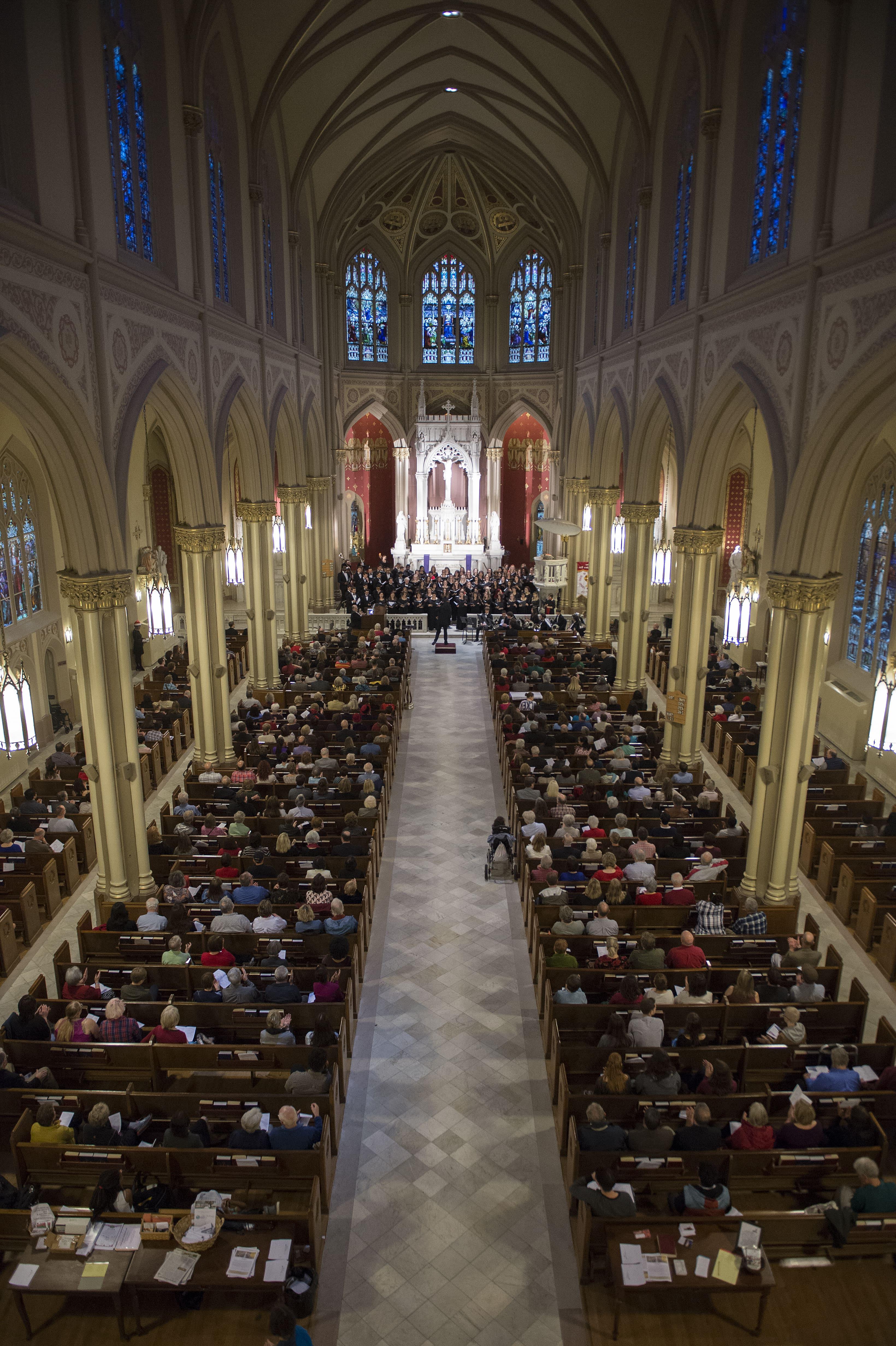 A high aerial shot of Holy Name of Jesus Church, filled with an audience listening to singers and instrumentalists performing on the altar.